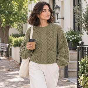 May include: An olive green long-sleeved jumper featuring a chunky cable knit pattern on the front and sleeves, a round neckline, and ribbed cuffs. The jumper is paired with light-coloured trousers and a plain beige canvas tote bag.