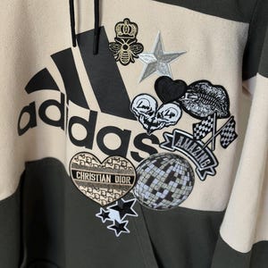 Op de afbeelding: Een groene, beige en witte Adidas hoodie met een groot Adidas logo. De hoodie is versierd met verschillende patches, waaronder een bij, een ster, een hart, lippen en een discobal. De tekst "CHRISTIAN DIOR" is ook zichtbaar.