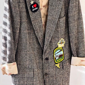 May include: A vintage plaid blazer in shades of gray and brown, featuring rolled-up sleeves with a patterned lining. The blazer is adorned with pins, including one that says "I ❤️ MUSIC" and a patch that says "City of Folsom R.T.C."