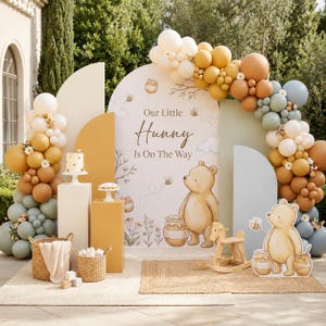 May include: A baby shower backdrop featuring a large arched sign with the text Our Little Hunny Is On The Way, illustrated Winnie the Pooh characters, honey pots, and bees, surrounded by a balloon garland in shades of cream, gold, and sage.