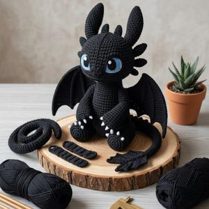 May include: A black crochet dragon with large blue eyes, wings, and white accents on its feet. The dragon is sitting on a wooden slice, with yarn and other crochet pieces nearby. A small potted succulent is in the background.