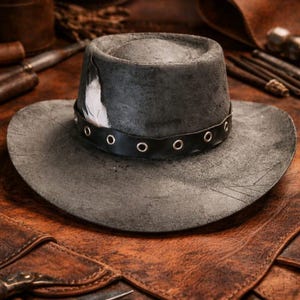 Peut inclure: Un chapeau gris anthracite à l'aspect vieilli, doté d'une calotte plate et d'un large bord. Il est orné d'une bande en cuir noir avec des œillets métalliques argentés et d'une plume blanche et noire insérée sur le côté de la bande.