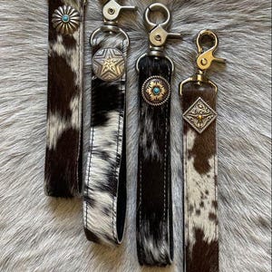 May include: Four keychains featuring brown and white cowhide leather straps attached to silver-toned metal swivel lobster clasps. Each strap is adorned with a unique decorative metal concho, including star and floral designs with turquoise accents.