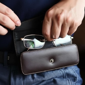 Könnte beinhalten: Eine Brille mit goldenem Rahmen wird aus einem braunen Lederetui entnommen. Das Etui, das mit einem Druckknopf verschlossen ist, wird von einem schwarzen Gürtel gehalten. Die Person trägt eine blaue Jeans und ein dunkelblaues Hemd.