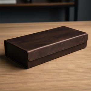 以下が含まれることがあります： 長方形の濃い茶色の革製ボックスで、ヒンジ付きの蓋が付いています。ボックスはテクスチャ加工された表面で、2つの円形のステッチのディテールで固定されています。ボックスは明るい茶色の木製の表面にあります。