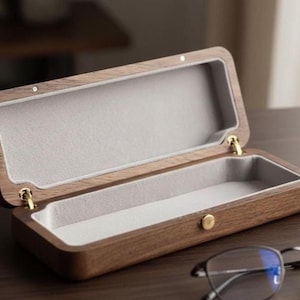 Könnte beinhalten: Geöffnete Brillenetui aus Holz mit grauem Filzinnenraum. Das Etui ist rechteckig mit abgerundeten Kanten und einem Messingverschluss. Die Scharniere sind ebenfalls aus Messing. Das Etui ist aus dunkelbraunem Holz.