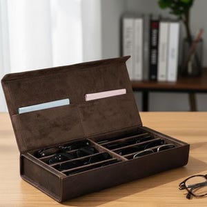 May include: A brown leatherette glasses case, open to reveal multiple compartments holding various pairs of eyeglasses. The interior is lined with a soft, dark brown material. Two cleaning cloths are tucked into the lid. A pair of glasses rests on the table.