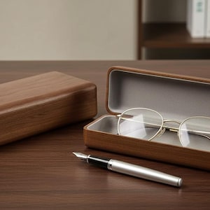 以下が含まれることがあります： 丸いフレームの眼鏡が入った木製の眼鏡ケース。閉じた木箱と銀色のペンも濃い木材の表面にあります。ケースの内側は明るいグレーです。