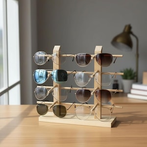 Peut inclure: Un présentoir en bois pour lunettes de soleil, présentant différentes paires avec des montures et des verres de couleurs variées. Le présentoir en bois clair comporte trois niveaux pour accueillir plusieurs paires. En arrière-plan, un bureau, une lampe et une fenêtre.