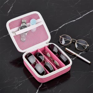 May include: A pink and white eyeglasses case with a zip, containing sunglasses and spectacles. A watch and contact lens case are in the zipped pocket. Gold-rimmed glasses and a pen are next to the case.