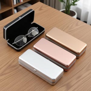 Könnte beinhalten: Ein Set aus vier rechteckigen Hartschalenetuis für Brillen, bestehend aus einem offenen schwarzen Etui mit einer silbernen Brille sowie drei geschlossenen Etuis in den Metallic-Farben Silber, Roségold und Gold mit glatter, einfarbiger Oberfläche.