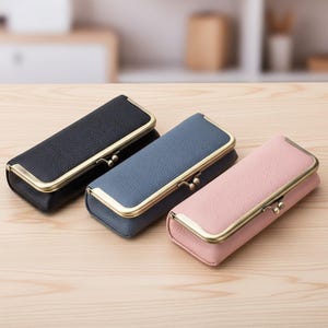 May include: Three rectangular cases in black, blue, and pink, each with a textured leather-like finish. The cases have gold-colored metal frames and clasps. The cases are arranged on a light-colored wooden surface.