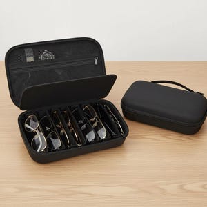 May include: Black sunglasses case with multiple compartments, holding various pairs of sunglasses. A separate, closed black case with a handle sits beside the open case. A watch is visible in the open case.