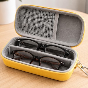 Puede incluir: Un estuche rígido rectangular de color amarillo brillante para gafas con forro interior gris. El estuche está abierto y muestra dos pares de gafas en compartimentos separados, con un bolsillo de malla en la tapa y un clip de metal.