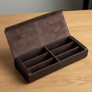 以下が含まれることがあります： 6つのコンパートメントを備えた、開いた茶色の革製メガネケース。内側は柔らかい濃い茶色の素材で裏打ちされています。ケースにはヒンジ付きの蓋があり、明るい木製の表面に置かれています。