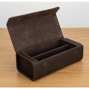 以下が含まれることがあります： ヒンジ付きの蓋が付いた茶色の革製の箱で、2つの内部コンパートメントが現れるように開いています。 箱は角が丸い長方形で、2つの円形のスナップ留めで固定されています。 内側は柔らかい濃い茶色の素材で裏打ちされています。