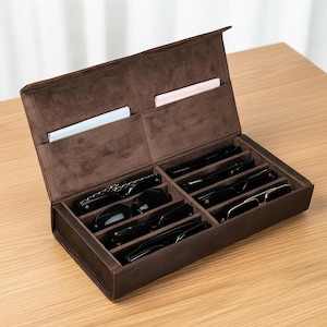 May include: A brown leather eyeglasses case, open to show compartments filled with sunglasses and spectacles. The interior is lined with a soft, dark brown material. Two small pockets hold a light blue and a pink cleaning cloth.