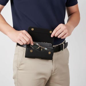 May include: A black rectangular belt pouch made of a textured material with a flap closure secured by two gold-toned metal snaps. The pouch is attached to a black leather belt and contains a smartphone and a pair of thin-framed metal glasses.