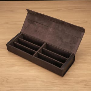 以下が含まれることがあります： 茶色の革製ペンケース。ヒンジ付きの蓋を開けると、複数のコンパートメントが現れます。内側は柔らかい濃い茶色の素材で裏打ちされています。ケースは長方形で、明るい木製の表面に置かれています。