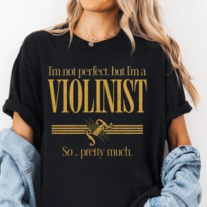May include: A black short-sleeve t-shirt featuring gold text that reads "I'm not perfect, but I'm a VIOLINIST So... pretty much." Below the text is a gold graphic of a violin and musical staff lines. The shirt is worn with a denim jacket.