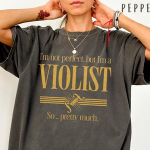 Könnte beinhalten: Ein anthrazitfarbenes Kurzarm-T-Shirt mit goldenem Text, der lautet: "I'm not perfect, but I'm a VIOLIST So... pretty much." Das Design enthält eine Grafik eines Violinstegs und Saiten, die zwischen den Textzeilen zentriert ist.