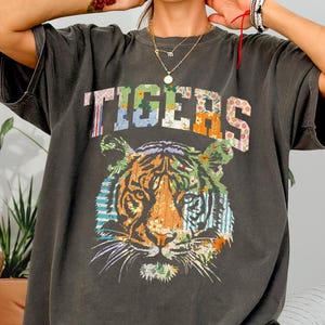 May include: A charcoal grey short-sleeved t-shirt featuring the word TIGERS in a multi-coloured floral and striped pattern across the chest. Below the text is a large graphic of a tiger head composed of various colourful floral and striped patches.
