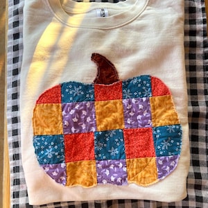 May include: A cream-colored crewneck sweatshirt featuring a large, central pumpkin applique. The pumpkin is constructed from a patchwork of quilted fabric squares in vibrant shades of orange, yellow, purple, and blue with floral patterns.
