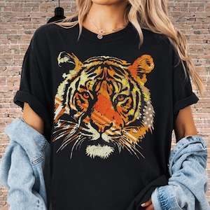 May include: A black short-sleeve t-shirt featuring a large, stylized graphic of a tiger face in shades of orange, yellow, and white. The tiger illustration includes diagonal line patterns and a dotted texture on the right side of the design.