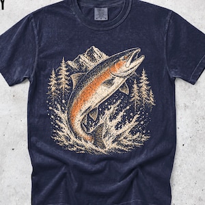 K&ouml;nnte beinhalten: Ein marineblaues Kurzarm-T-Shirt mit einem Grafikdruck eines springenden Fisches in Orange- und Cremet&ouml;nen. Die Illustration zeigt eine Bergkette und Kiefern im Hintergrund sowie Wasser, das um den Fisch spritzt. Das Wort NAVY ist in Schwarz in der oberen linken Ecke aufgedruckt.