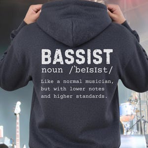 May include: A dark charcoal grey hooded sweatshirt featuring white text on the back that reads: BASSIST noun /ˈbeɪsɪst/ Like a normal musician, but with lower notes and higher standards. The hoodie has long sleeves, a hood, and a ribbed hem.