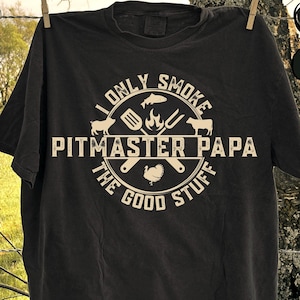 May include: Black t-shirt with white text and graphics. The text reads "I Only Smoke Pitmaster Papa The Good Stuff." The design includes images of a pig, fish, fire, and turkey. The shirt is hanging on a clothesline.