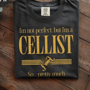 May include: A black crew neck t-shirt featuring gold text that reads I'm not perfect, but I'm a CELLIST So... pretty much. Below the text is a gold graphic of a cello scroll and neck positioned over horizontal musical staff lines.