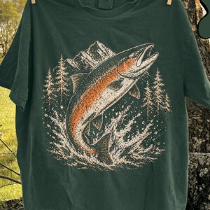 K&ouml;nnte beinhalten: Ein dunkelgr&uuml;nes Kurzarm-T-Shirt mit einem Grafikdruck eines aus dem Wasser springenden Fisches vor einem Hintergrund aus Bergen und Tannen. Das Motiv verwendet Orange-, Wei&szlig;- und Schwarzt&ouml;ne. Der Text BLUE SPRUCE ist deutlich zu sehen.