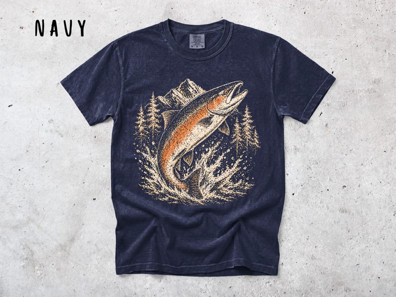K&ouml;nnte beinhalten: Ein marineblaues Kurzarm-T-Shirt mit einem Grafikdruck eines springenden Fisches in Orange- und Cremet&ouml;nen. Die Illustration zeigt eine Bergkette und Kiefern im Hintergrund sowie Wasser, das um den Fisch spritzt. Das Wort NAVY ist in Schwarz in der oberen linken Ecke aufgedruckt.