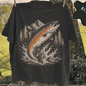 K&ouml;nnte beinhalten: Ein schwarzes T-Shirt mit einer grafischen Illustration eines Fisches, der aus dem Wasser springt. Das Design zeigt im Hintergrund ein Gebirge und Tannen in Orange-, Wei&szlig;- und Schwarzt&ouml;nen. Das Wort BLACK ist in wei&szlig;er Schrift aufgedruckt.