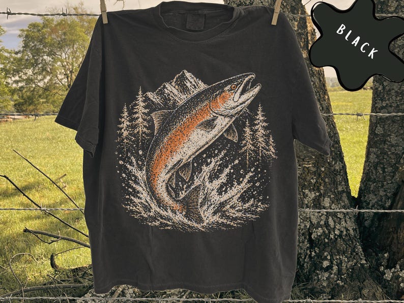 K&ouml;nnte beinhalten: Ein schwarzes T-Shirt mit einer grafischen Illustration eines Fisches, der aus dem Wasser springt. Das Design zeigt im Hintergrund ein Gebirge und Tannen in Orange-, Wei&szlig;- und Schwarzt&ouml;nen. Das Wort BLACK ist in wei&szlig;er Schrift aufgedruckt.