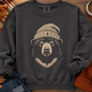 May include: A charcoal gray crewneck sweatshirt featuring a graphic illustration of a black bear wearing a beige knit beanie. The bear is centered on the chest, depicted with a light-colored chest patch and a small pinecone icon on the beanie.