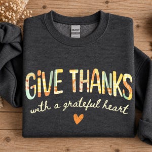 May include: A dark charcoal grey crewneck sweatshirt featuring the text "GIVE THANKS with a grateful heart" in a multi-coloured, patterned font with autumn-inspired hues of orange, yellow, and teal, accompanied by a small orange heart graphic.