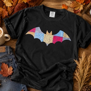 May include: A black short-sleeved t-shirt featuring a central bat graphic composed of a colourful patchwork pattern in shades of blue, yellow, and red. The shirt has a crew neckline and a Comfort Colors brand label printed on the inside collar.