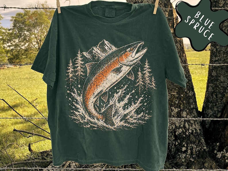 K&ouml;nnte beinhalten: Ein dunkelgr&uuml;nes Kurzarm-T-Shirt mit einem Grafikdruck eines aus dem Wasser springenden Fisches vor einem Hintergrund aus Bergen und Tannen. Das Motiv verwendet Orange-, Wei&szlig;- und Schwarzt&ouml;ne. Der Text BLUE SPRUCE ist deutlich zu sehen.