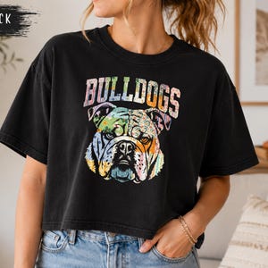 May include: A black short-sleeved t-shirt featuring a graphic of a bulldog head with a multi-colored, patterned design. The word BULLDOGS is printed in a large, arched, multi-colored font above the dog illustration. The shirt has a crew neckline.