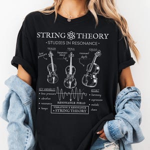 May include: A black t-shirt featuring a white graphic print titled STRING THEORY STUDIES IN RESONANCE. The design displays line drawings of a violin, viola, and cello with labels, a wave diagram labelled RESONANCE FIELD, and text equations.