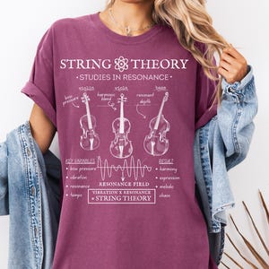 May include: A maroon t-shirt featuring a white graphic print titled String Theory Studies in Resonance. The design includes line drawings of a violin, viola, and bass, along with text labels for key variables, a resonance field wave, and the equation Vibration x Resonance = String Theory.