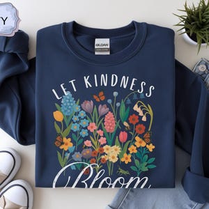 May include: Navy blue sweatshirt with a floral design and the words "Let Kindness Bloom" in white script. The design features colorful flowers and butterflies. The sweatshirt is paired with jeans and sneakers.