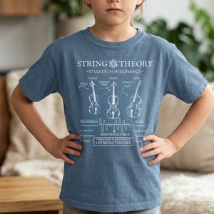 May include: A dusty blue short-sleeve t-shirt featuring a white graphic print titled STRING THEORY STUDIES IN RESONANCE. The design includes line drawings of a violin, viola, and bass, along with scientific text labels and a wave diagram.
