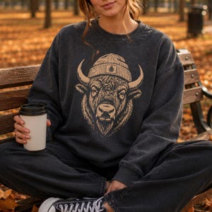 May include: A charcoal gray crewneck sweatshirt featuring a tan graphic illustration of a bison wearing a beanie. The bison is centered on the chest, depicted with detailed line work, prominent horns, and a small pinecone icon on the beanie.