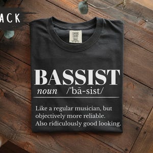 May include: A black t-shirt featuring white text that reads BASSIST noun /'ba-sist/ Like a regular musician, but objectively more reliable. Also ridiculously good looking. The shirt has a crew neckline and a Comfort Colors brand label at the neck.