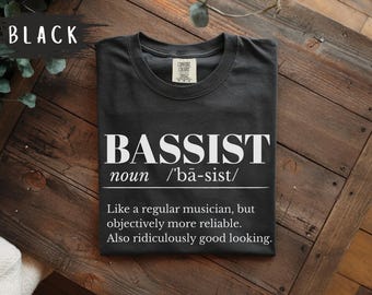 May include: A black t-shirt featuring white text that reads BASSIST noun /'ba-sist/ Like a regular musician, but objectively more reliable. Also ridiculously good looking. The shirt has a crew neckline and a Comfort Colors brand label at the neck.