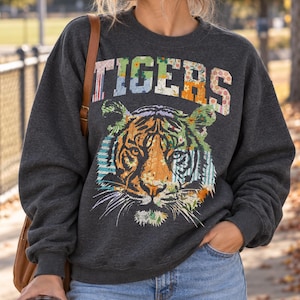 May include: A dark charcoal grey crewneck sweatshirt featuring the word TIGERS in a multi-coloured floral and striped patchwork font above a large, detailed tiger head graphic composed of various patterned fabric textures and colours.