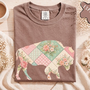 May include: Espresso-colored t-shirt featuring a floral patchwork buffalo design. The buffalo silhouette is filled with pastel floral patterns and plaid squares. The shirt is displayed on a wooden surface with a cup of coffee and floral accents.